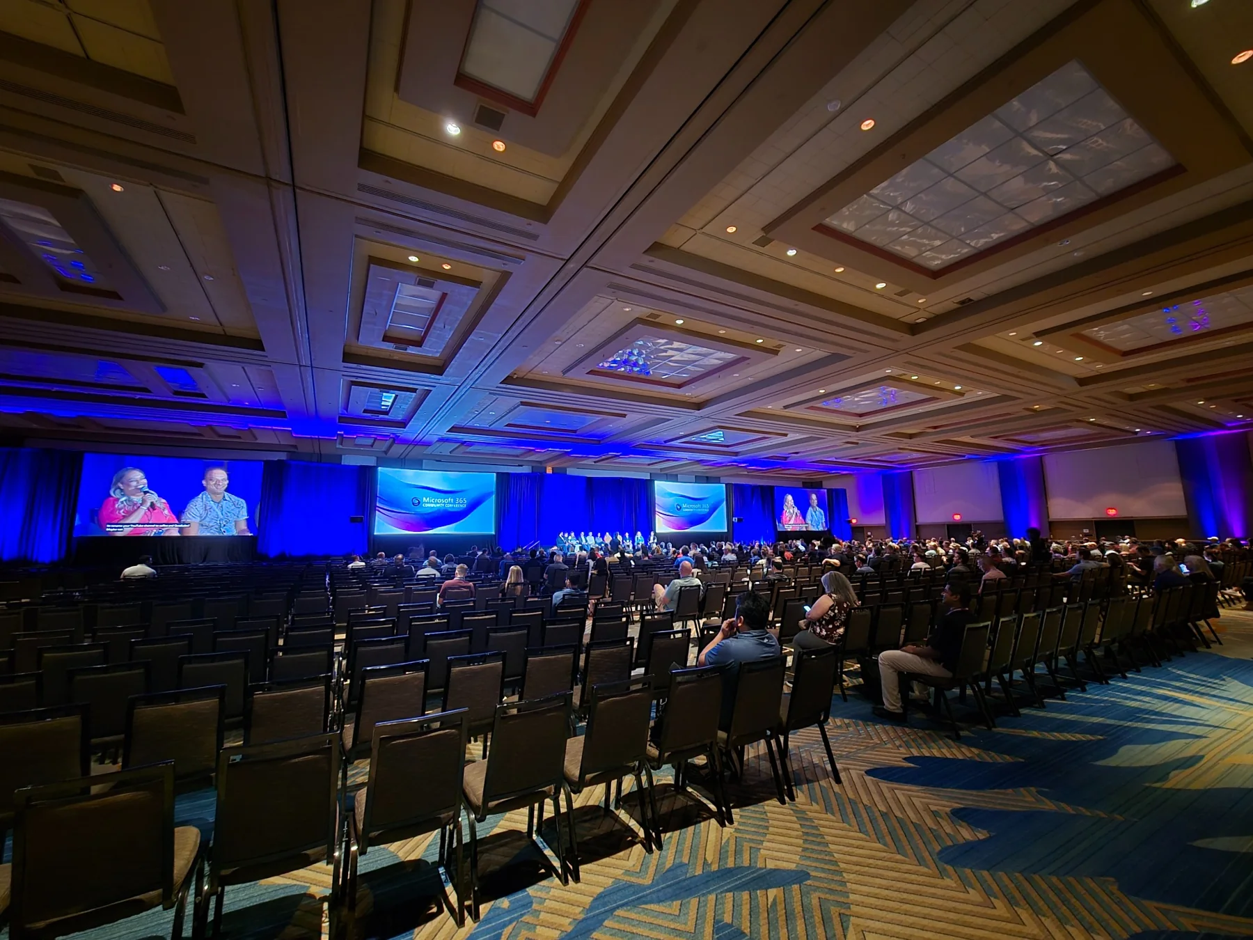Wide corporate conference ballroom with blue stage lighting, multiple large screens, and audience seating