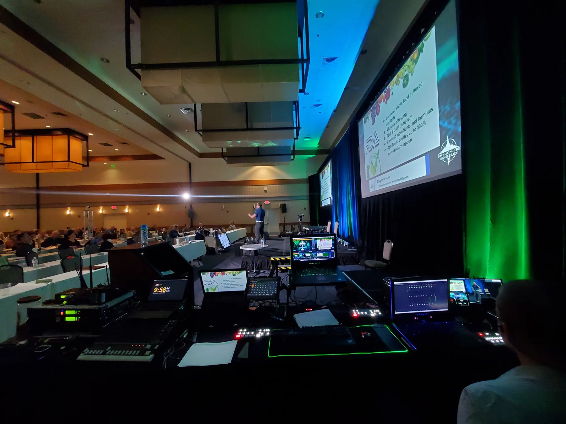 Front-of-house control position looking toward a live corporate stage with switching, playback, and lighting control visible