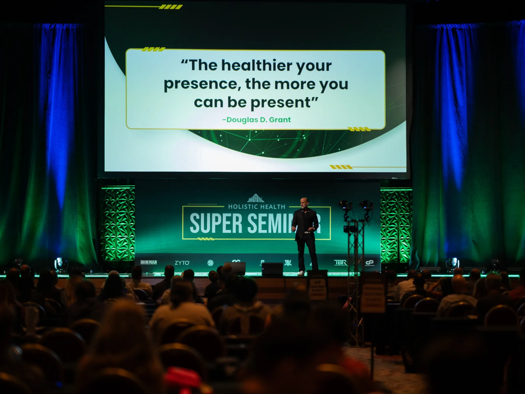 Keynote speaker on a large conference stage beneath a quote slide and branded event backdrop