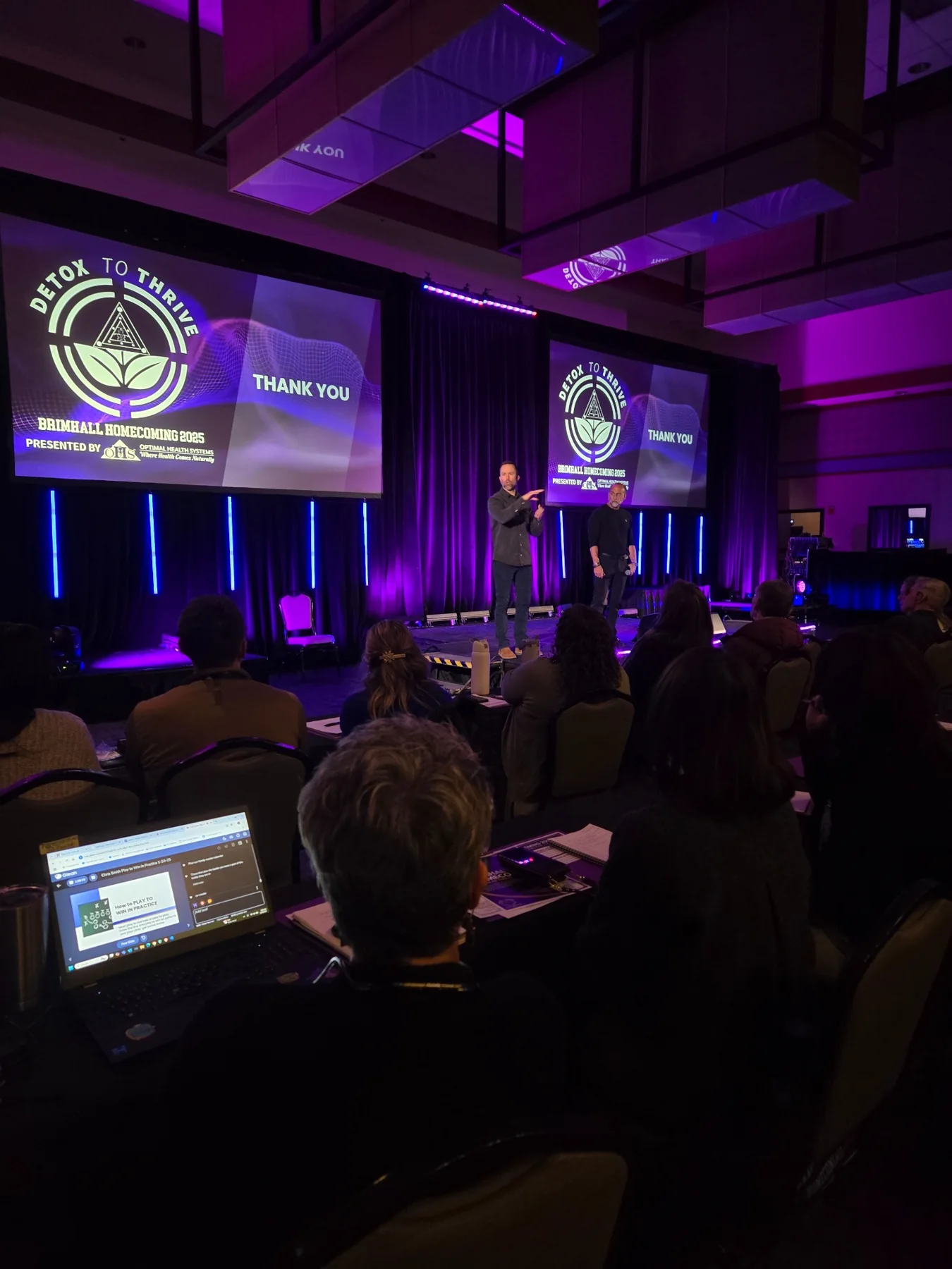Audience seated at tables during a live corporate session with purple lighting and twin presentation screens