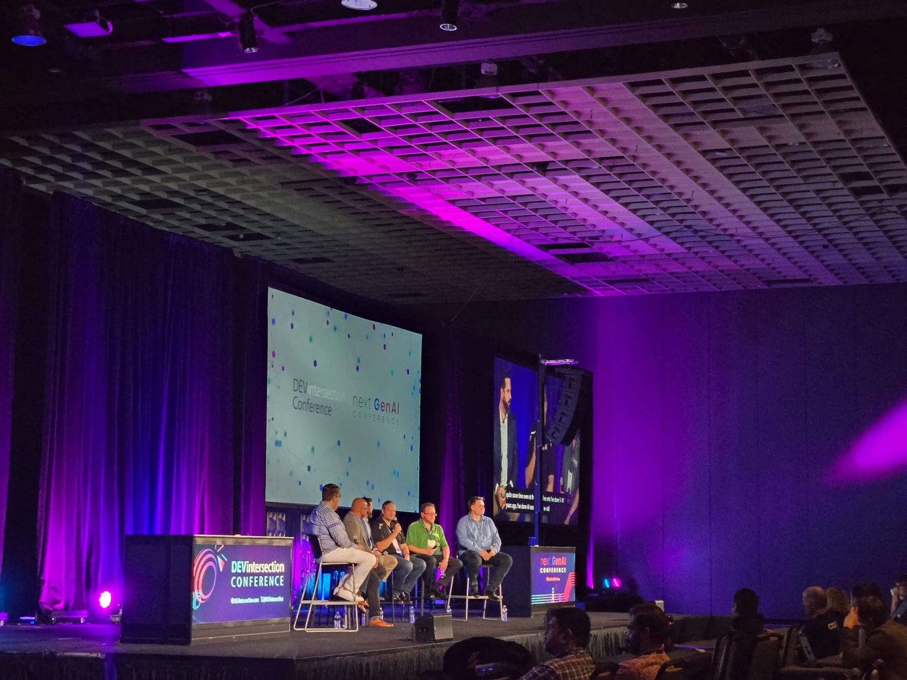 Corporate panel discussion on stage with branded side screens and purple show lighting