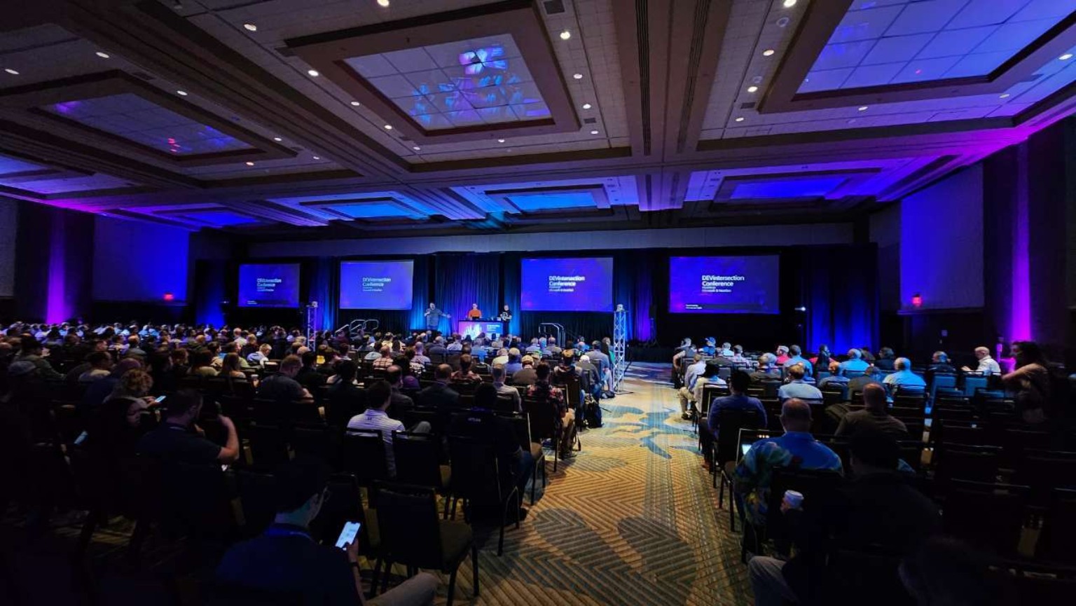 Wide ballroom view with a full audience, multiple projection screens, and coordinated blue and purple room lighting