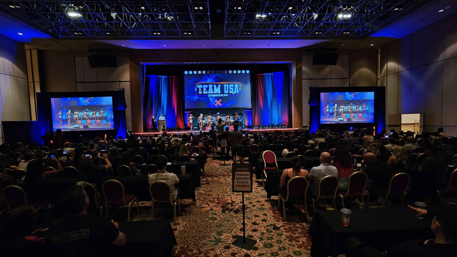 Live stage production with a centered screen, two side screens, blue lighting, and a seated audience