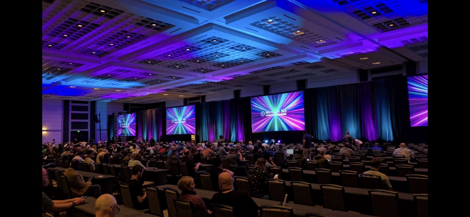 Full M365 conference room with colorful presentation screens, blue and purple ceiling lighting, and a seated audience